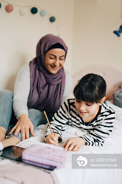 Smiling mother looking at daughter writing in book on bed at home