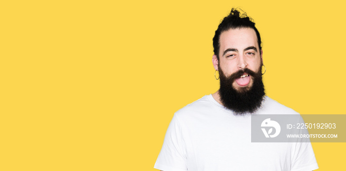 Young hipster man with long hair and beard wearing casual white t-shirt sticking tongue out happy wi