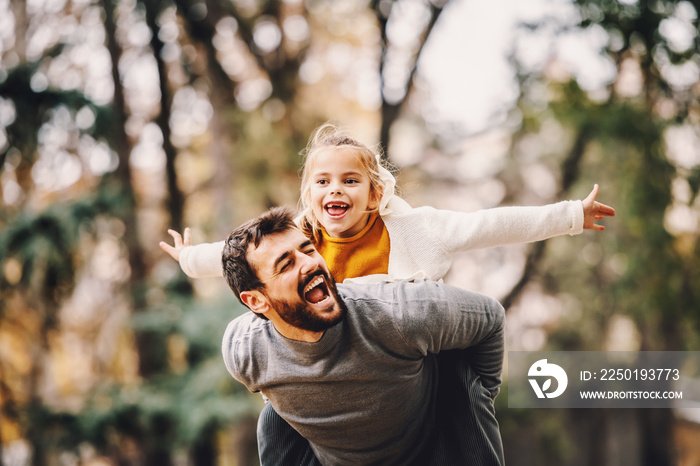 Playful handsome bearded Caucasian father having piggyback with his adorable daughter. Autumn in par