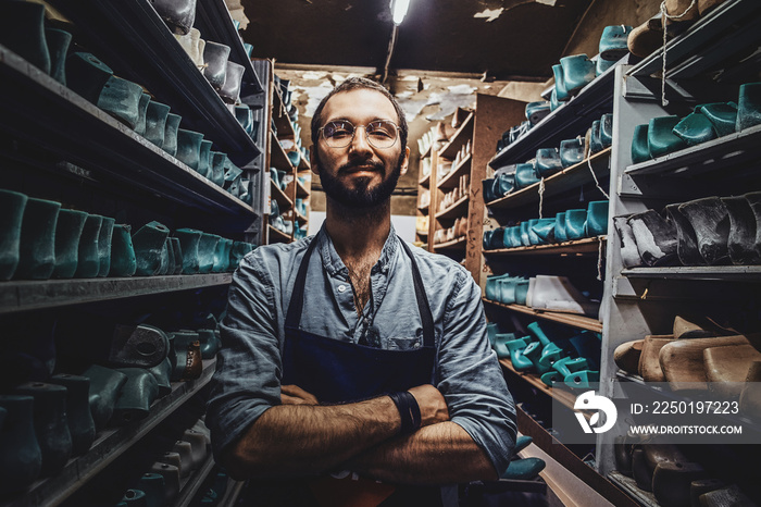 Bearded young cobbler in glasses is posing for photographer at dark shoe form storage.