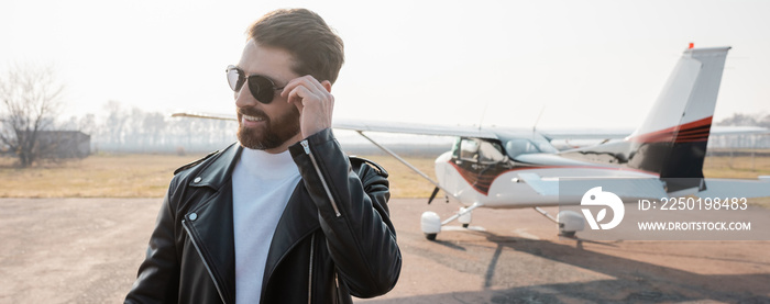 pleased pilot in stylish leather jacket adjusting sunglasses near helicopter, banner.