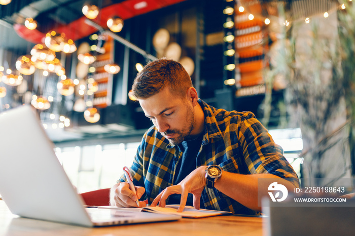 Focused bearded Caucasian freelancer writing in notebook tasks while sitting in cafeteria. On table 