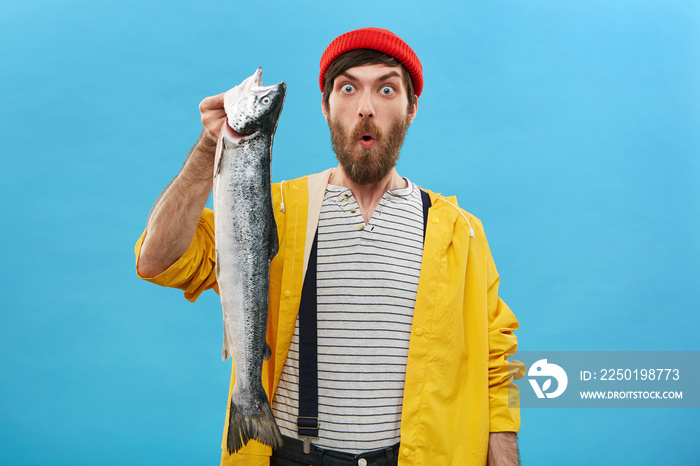 Attractive male with beard dressed in red hat, yellow raincoat and overalls holding huge fish lookin