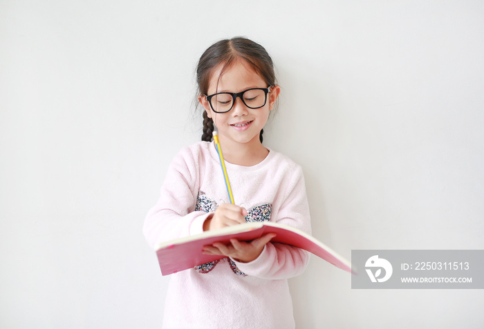 快乐的亚洲小女孩戴着眼镜，手里拿着书，在白背上用铅笔写字