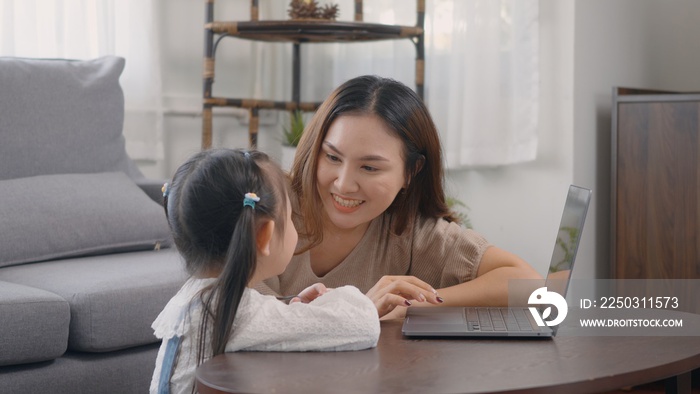 Asian young mother with laptop computer teaching her kid to learn or study online in living room at 