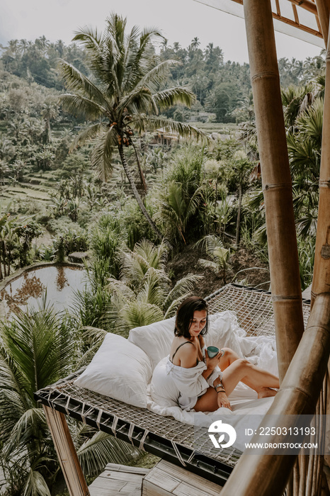 A young travelling woman relaxing in the lounge area of a Bali jungle hotel surrounded by jungle, pa