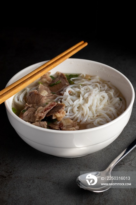 Vertical shot of soup Pho Bo with sticks on it and with a spoon on the side