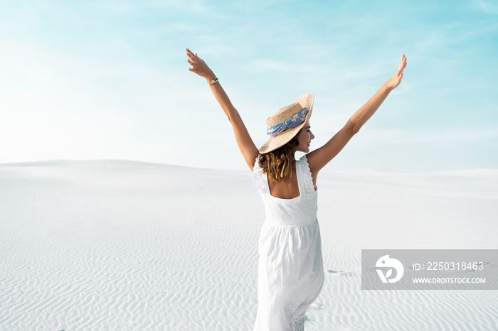 back view of beautiful girl in white dress and straw hat on sandy beach with blue sky