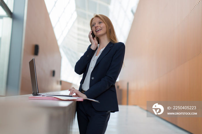 Businesswoman talking on smart phone at laptop in modern office