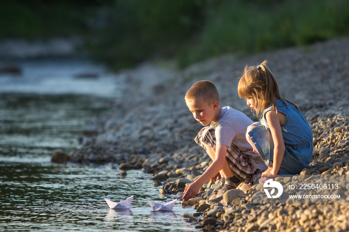 Two cute blond children, boy and girl on river bank sending in water white paper boats on bright sum