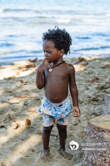 Imagen vertical de un pequeño niño afroamericano de cabello afro de pie solo en la playa con su dedo