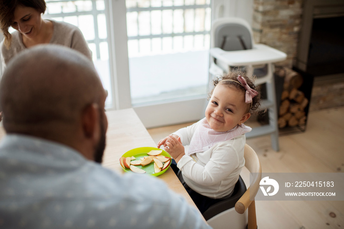 High angle portrait of cute daughter eating apple while sitting with parents at dining table