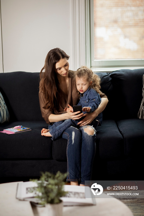 Mother with cute daughter using smart phone while sitting on sofa against wall at home