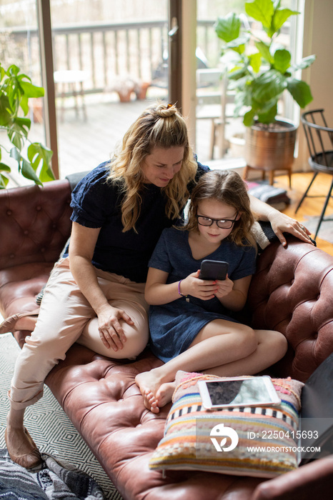 High angle view of mother looking at cute daughter using mobile phone while sitting on sofa in livin