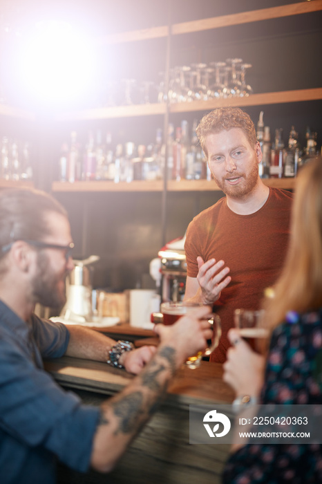 Cheerful friends leaning on bar counter, drinking beer and chatting with bartender. Night out.