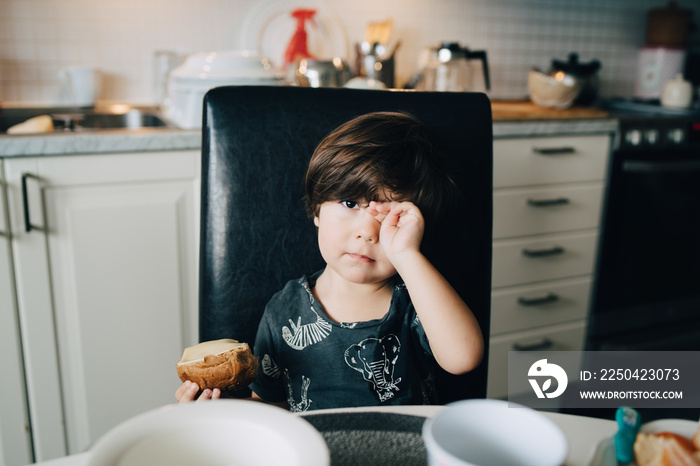 Cute boy eating bread while sitting at dining table in kitchen