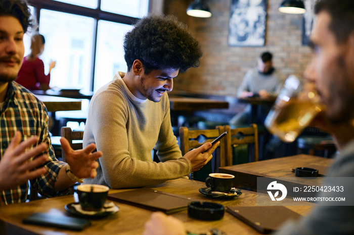 Happy Lebanese man texting on cell phone while being with his friends in cafe,