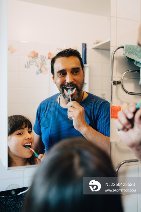 Father and daughter brushing teeth while looking at mirror in bathroom