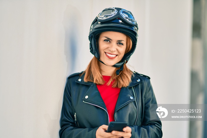 年轻的拉丁女子戴着摩托车头盔在城市里使用智能手机。