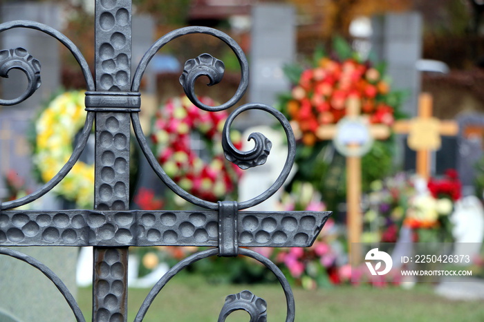 Am Friedhof, Im Vordergrund ein schmiedeeisernes Grabkreuz, im unscharfen Hintergrund Blumenkränze a