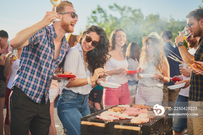 Group of people standing around grill, chatting, drinking and eating.