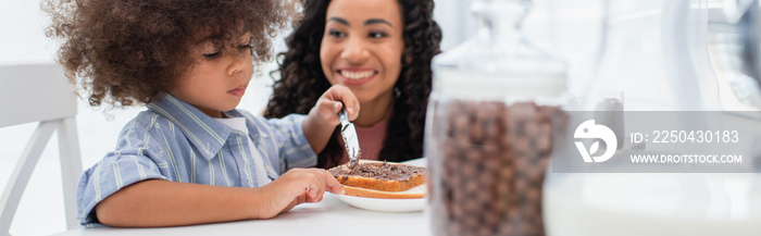 积极的非裔美国妈妈看着蹒跚学步的女儿在面包上涂巧克力酱，横幅