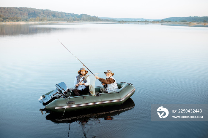 Grandfather with adult son fishing on the inflatable boat on the lake with calm water early in the m