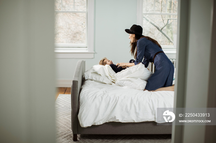 Side view of playful mother wearing hat tickling daughter lying on bed at home seen through doorway