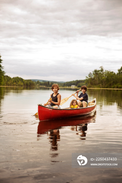 Boys (12-13, 8-9) and girl (4-5) on lake in red boat, looking at camera trees in background