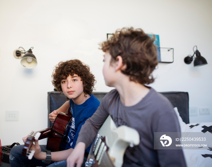 Brothers practicing guitars while sitting on bed against wall at home