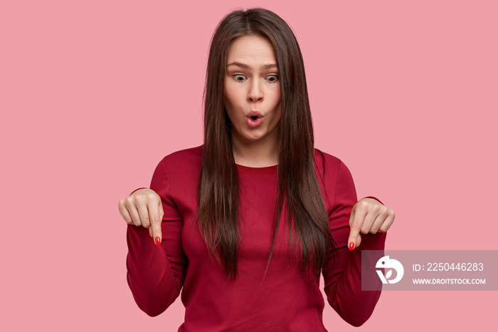 Emotional brunette young Asian woman points down with both index finger on floor, opens mouth from s