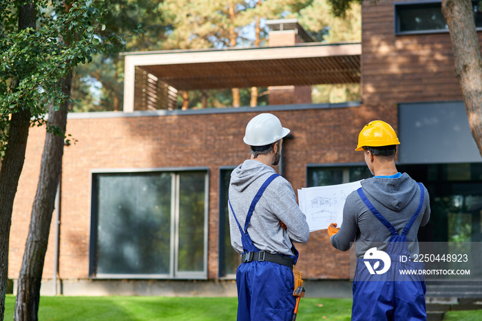 Making the best house. Rear view of two builders standing outdoors with an open blueprint discussing