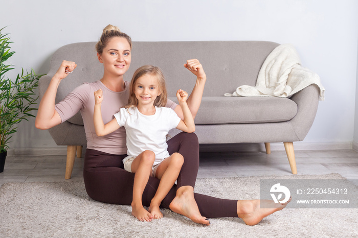 Attractive fit mom and her charming daughter showing biceps, smiling at camera, sitting together on 