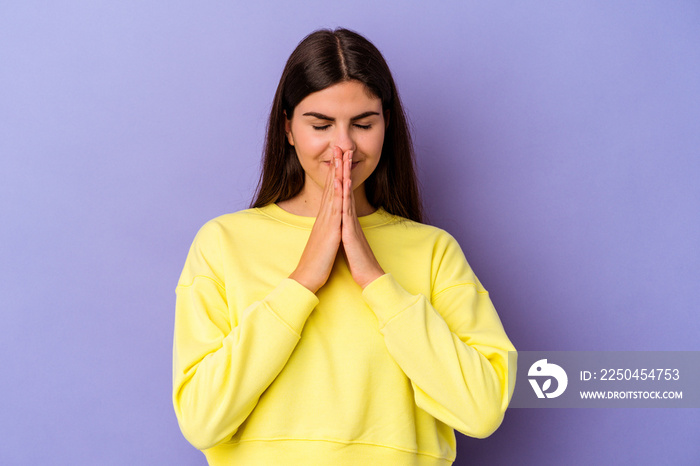 Young caucasian woman isolated on purple background holding hands in pray near mouth, feels confiden