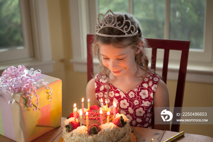 Girl with crown looking at birthday cake while sitting on chair at home