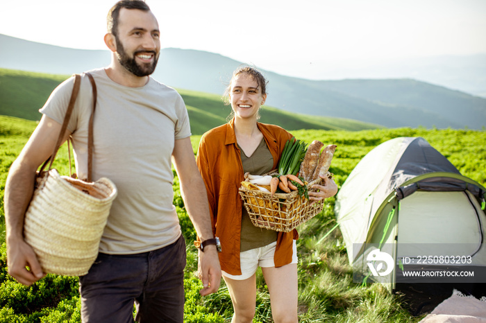 Young and cheerful couple walking near the campsite in the mountains, carrying basket full of fresh 