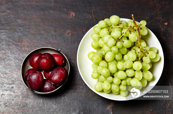 Branch of ripe green grape on plate with water drops and plums in bowl. Juicy fruits on wooden backg