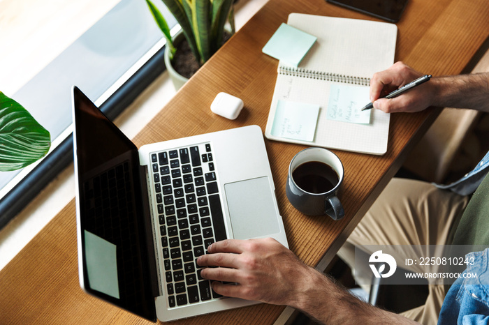 Photo of man writing notes and using laptop while working in cafe indoors