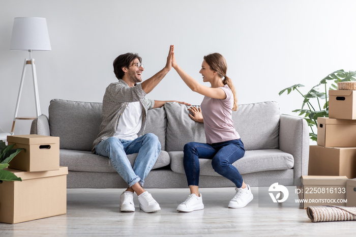 Satisfied european young husband and wife give high five on sofa in living room interior with boxes