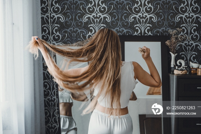 How take care of long hair. Back view of young natural blonde woman in white home clothes combing he