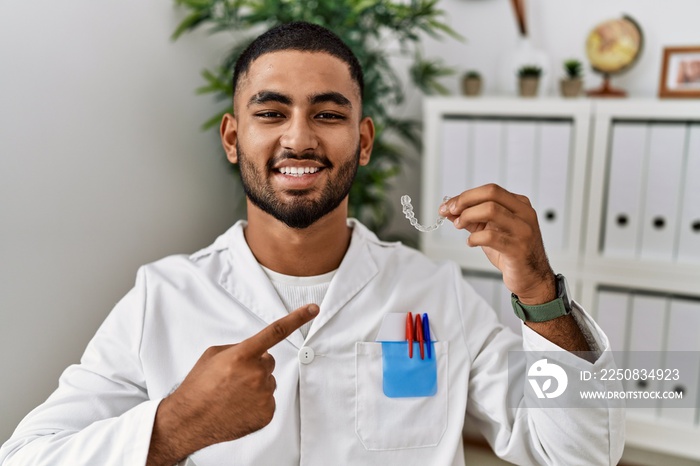 Young indian man working at dentist clinic holding invisible aligner smiling happy pointing with han