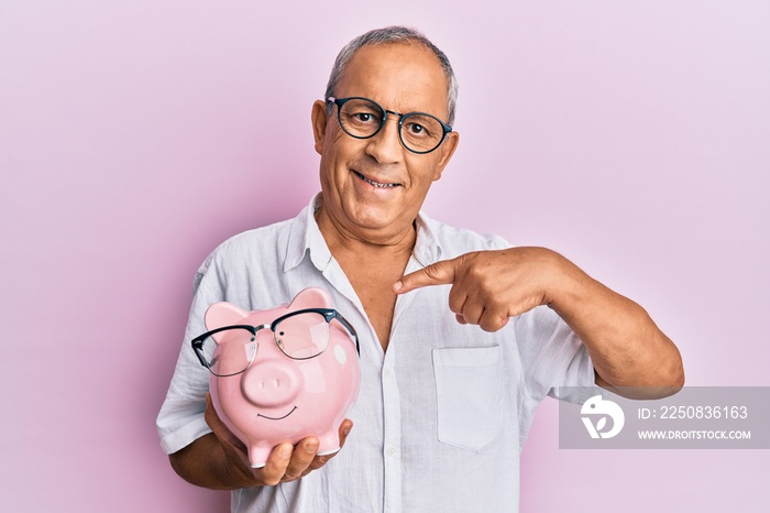 Handsome mature senior man holding piggy bank with glasses smiling happy pointing with hand and fing