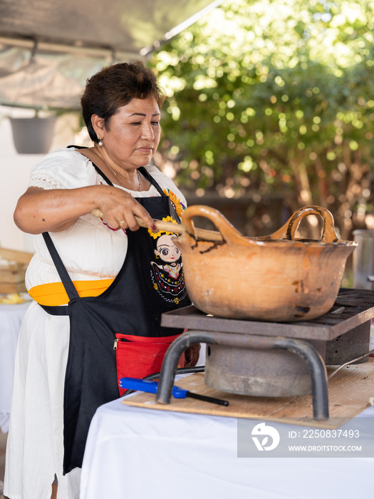 Wide-angel vertical image of an Indigenous woman stirring mole in a clay pot with a big wooden spoon