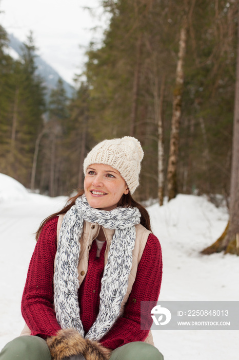 Portrait smiling woman in knit hat and scarf sitting in snowy woods