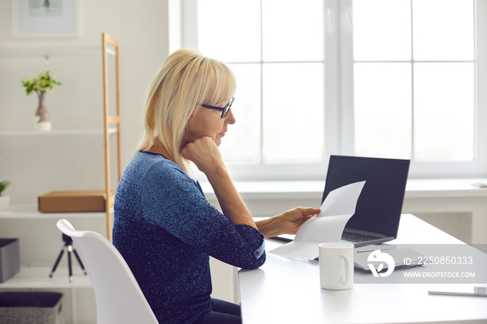 Side view unhappy older adult female reading paper sheet document, sitting at desk at home. Sad reti