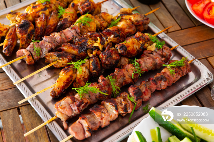Appetizing shashliks of pork meat and chicken grilled on skewers served with fresh cucumbers and gre