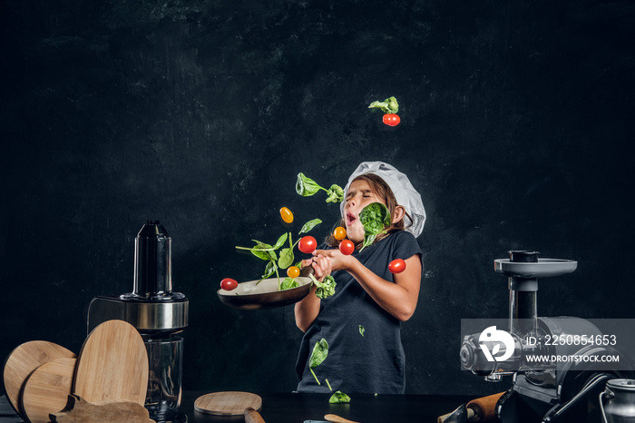 Funny little girl is tossing vegetables on the pan at dark photo studio.