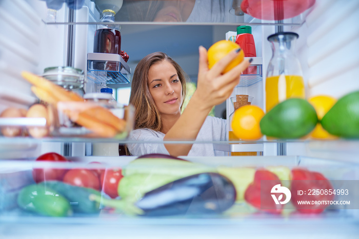 一个女人站在打开的冰箱前拿着橙色水果。冰箱里装满了杂货。图片