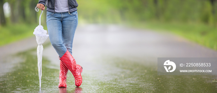 Detail of female legs in rain boots and a closed umbrella standing on the road on a rainy day