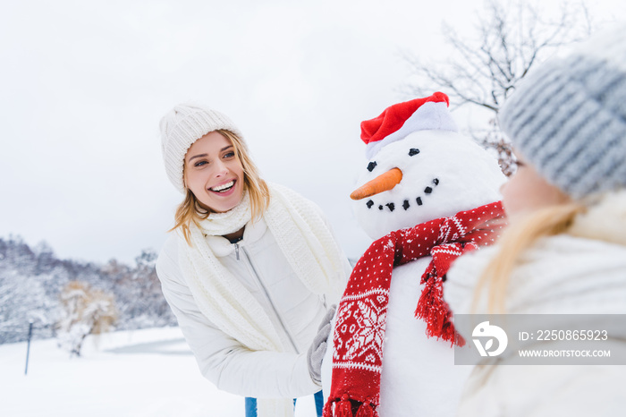 happy mother and daughter standing near snowman together in winter forest